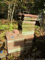 Stack of concrete square pavers with some moss and broken pieces on top outdoors among foliage.