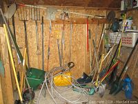 Wide view of shed wall showing an assortment of yard and garden tools including several snow shovels, rakes, pitch forks, and a garden spreader.