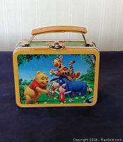 Front view of tin lunch box showing colorful image of Pooh, Tigger, Piglet, and Eeyore in a flower field scene with yellow border and handle.