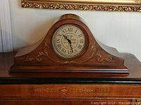 Front view of the wooden mantle clock showing ornate inlaid decoration and round clock face with Roman numerals.