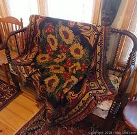 Full view of throw or rug draped over wooden furniture showing sunflower and floral pattern with scroll border and fringed edges.
