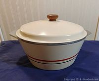 Front-side angle view of off-white cast iron Dutch oven with lid, wooden knob, and red/gray decorative stripes.