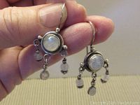 Photo of the front of the earrings held in hand showing central round moonstone cabochon and dangling oval and round moonstones with sterling silver findings.