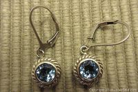 Front view of the pair of sterling silver earrings featuring round faceted blue topaz stones set in twisted rope pattern bezels with lever-back closures, laid out on a textured surface.
