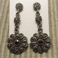 Front view of dangling sterling silver earrings with floral motif, marcasite stones, and central garnet coloured stone.