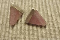 Front view of two triangular rhodochrosite stone earrings in sterling silver settings with a soft pink color on a textured beige fabric background.
