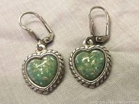 Front view of both sterling silver earrings with heart-shaped green lucite cabs with embedded specks and twisted rope silver border, resting on fabric.
