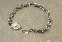 Braided sterling silver bracelet laid flat on a textured surface, showing full length and oval hook clasp with engraved letters GE.