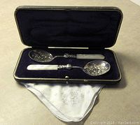 Two sterling silver serving spoons with mother of pearl handles inside an open purple velvet-lined presentation box with white embroidered cloth visible.