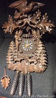 Front view of large carved wooden cuckoo clock showing bird on top, clock face with Roman numerals, forest animals and trees carved below, pinecone weights and leaf pendulum on the table in front.