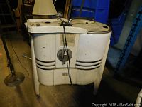 Side view of antique washing machine showing white metal body with black stripes, top compartments with lids, and attached faucet and wringer.