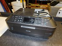 Front-side view of black Canon multifunction printer with keypad, control buttons, LCD screen, and power cord on top.