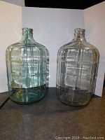 Two large glass jugs side by side against a white backdrop showing full body and neck.