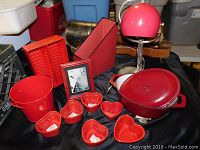 Photo showing large red enamelled cooking pot with lid, red plastic bucket, organizer, magazine file holder, six red heart-shaped ceramic dishes, vintage lamp, and framed photo on black cloth.