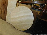 Photo of a round beige stone table top leaning against a wall, showing natural horizontal striations and smooth surface.