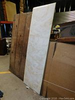 Full view of beige and white swirl marble countertop piece leaning against wall surrounded by boxes and wood planks.