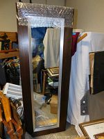 Floor length mirror with brown outer and silver inner wood frame, standing vertically against a cluttered background. The mirror surface is clear.
