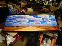 Wide panoramic framed road landscape artwork displayed on a table among other items.