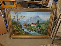 Full frontal view of oil painting in carved gold frame showing mountains, river and cottage