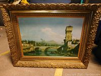 Frontal view of the framed painting showing a historical stone bridge and tower with an ornate gold frame.