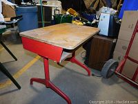 Front and side view of the small vintage school desk with red metal base and wooden desktop with visible wear.
