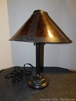 Unique 20-inch tall table lamp with riveted metal conical shade and dark metal base, placed on a dark surface with white backdrop.
