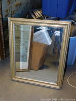 Rectangular gold framed mirror leaning against a wall with background storage items.