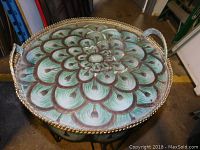Top view showing painted floral pattern on round metal tabletop with scalloped edges and rope-like trim. Two curved handles visible on opposite sides.