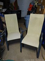 Two beige stacking patio chairs with black frames are photographed indoors in a garage-like setting with various storage containers and items around them.