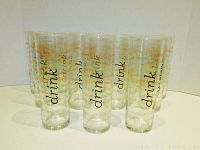 Front view of 12 clear tall drinking glasses with retro printed text and colorful designs arranged in a line.