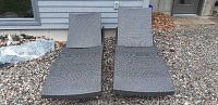 Two gray woven wicker/rattan chaise lounges side by side on gravel, viewed from front.