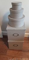 Stacked view of 5 grey storage boxes: three round on top of two square beneath on hardwood floor