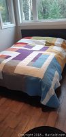 Full view of the quilt covering a queen-sized bed, showing color blocks in orange, purple, white, blue, and beige with a stitched texture.