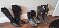Three pairs of ladies boots displayed side by side against a white wall on wooden floor. Includes black leather boots, black lizard print western boots with embroidery, and blue patterned rubber Bogs boots.