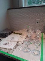 Overview of six crystal wine glasses, two smaller antique pressed glass champagne glasses, and assorted folded fabric placemats, napkins, and tea towels on kitchen counter.
