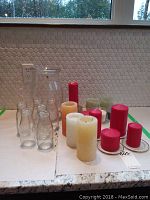 Photo showing red and white candles of various heights, glass milk jars, and a large clear glass vase on countertop.