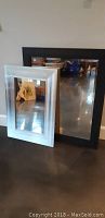 Two framed mirrors standing on floor side by side, one silver 20x28 and one black 27x33