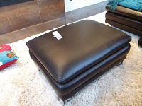 Top downward angled view showing black leather top cushion and overall rectangular shape of the ottoman, photo also captures some carpet and adjacent furniture.