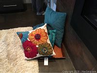 Three cushions stacked near wall, one with large floral embroidery in pink, orange, blue, and green hues, two teal cushions behind