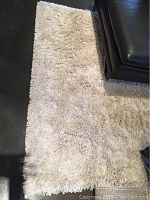 Top view of the cream-colored shaggy GÅSER area rug partially under a leather couch on a dark floor.