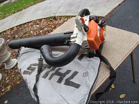 Full view of the Stihl SH85C gas powered leaf mulcher with attached collection bag and shoulder strap laid on table outdoors.