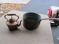 Two copper items on brown table surface: a wide copper pot and a smaller copper tea kettle with a curved handle and spout.