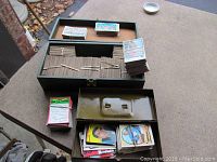 Overview of the sports cards collection showing an olive green metal box containing multiple bundles of cards, some loose stacks and small piles around it on an outdoor table.