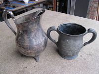 Image shows two silverplate vessels on a table; one is a rounded pitcher with a spout and the other a two-handled cup trophy with engraved writing.
