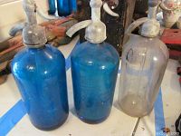 Three vintage seltzer bottles shown including two blue and one clear, all with metal siphon tops on a table.