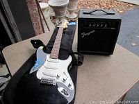 Black Fender Squier Stratocaster electric guitar with white pickguard and blue decals shown on a table alongside black padded guitar case and Traynor amplifier.