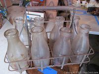 Set of 8 glass milk bottles arranged in original metal carrier showing general wear and rust