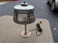 Side angle of Arts and Crafts Aladdin boudoir lamp showing painted frosted glass shade with floral and etched diamond pattern and round metal base with noticeable light pink chipping.