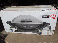 Boxed Weber Q portable propane grill, showing front and side views of packaging with images of the product and Weber branding.