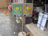 Photo showing both vintage ball point pen vending machines mounted on a single pole outside near a garage. The left machine has a green face, the right machine has a yellow face.